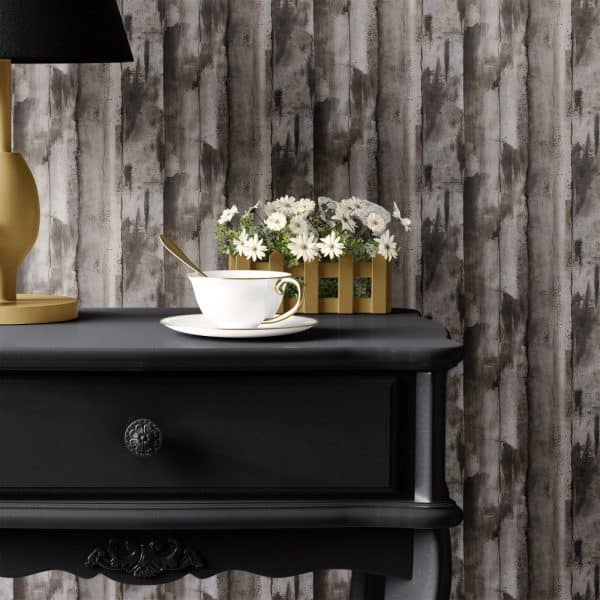 A White Teacup And Saucer Sit On A Black Side Table In Front Of A Wood-Grain Textured Wall. A Small Planter Of White Flowers Rests Beside The Teacup.