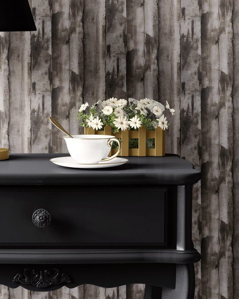 A white teacup and saucer sit on a black side table in front of a wood-grain textured wall. A small planter of white flowers rests beside the teacup.
