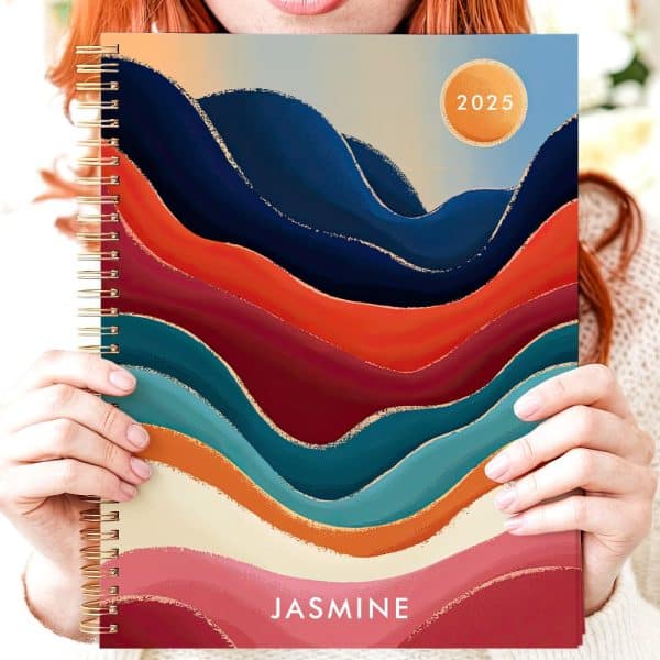 Person Holding A 2025 Spiral-Bound Planner With An Abstract Wave Design In Sunset Colors And Gold Accents. The Name Jasmine Is Printed On The Bottom.