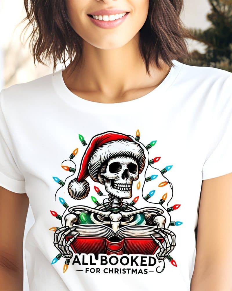 White t-shirt featuring a skeleton wearing a Santa hat and reading a book, surrounded by Christmas lights. The text "All Booked For Christmas" is below the image.