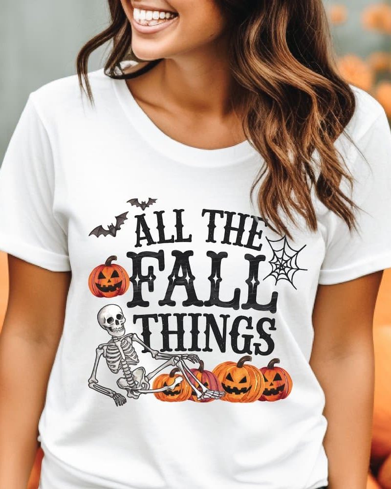 Woman wearing a white t-shirt with a Halloween design featuring a skeleton, pumpkins, bats, and a spiderweb. The shirt says All the Fall Things.