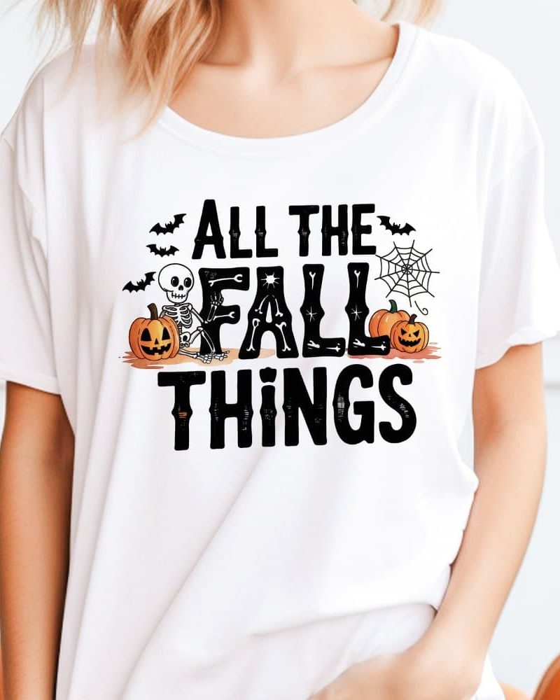White t-shirt with a Halloween-themed design. The design features a skeleton sitting between pumpkins, bats, a spiderweb, and the text All the Fall Things.