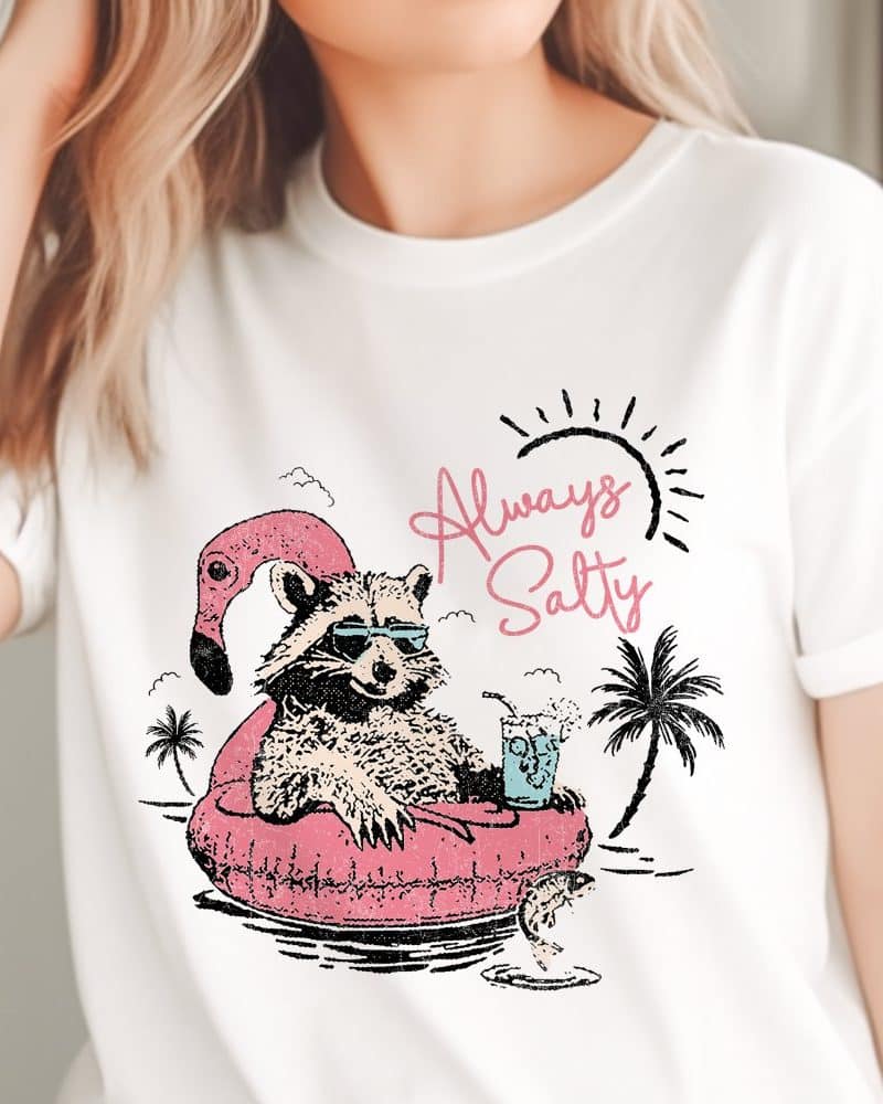 White t-shirt featuring a raccoon wearing sunglasses and relaxing on a pink flamingo floatie in the ocean. Text on the shirt reads Always Salty.