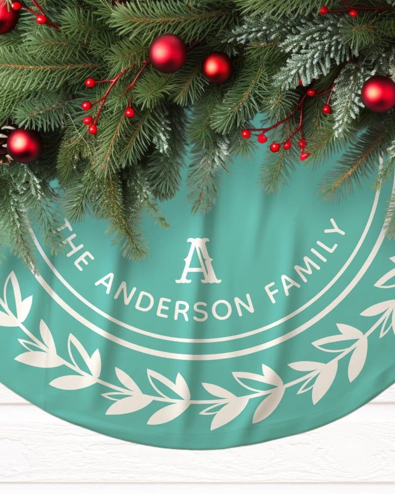 Teal Christmas tree skirt with the Anderson family name and a large A in a circular design, decorated with a wreath of leaves. Pine boughs with red ornaments are arranged at the top.