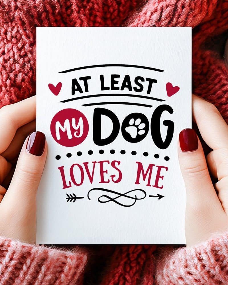 Two hands hold a card that says At Least My Dog Loves Me.