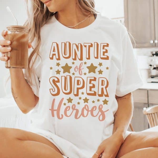 Woman Wearing A White T-Shirt That Says Auntie Of Super Heroes In Orange And Pink Lettering With Gold Stars.