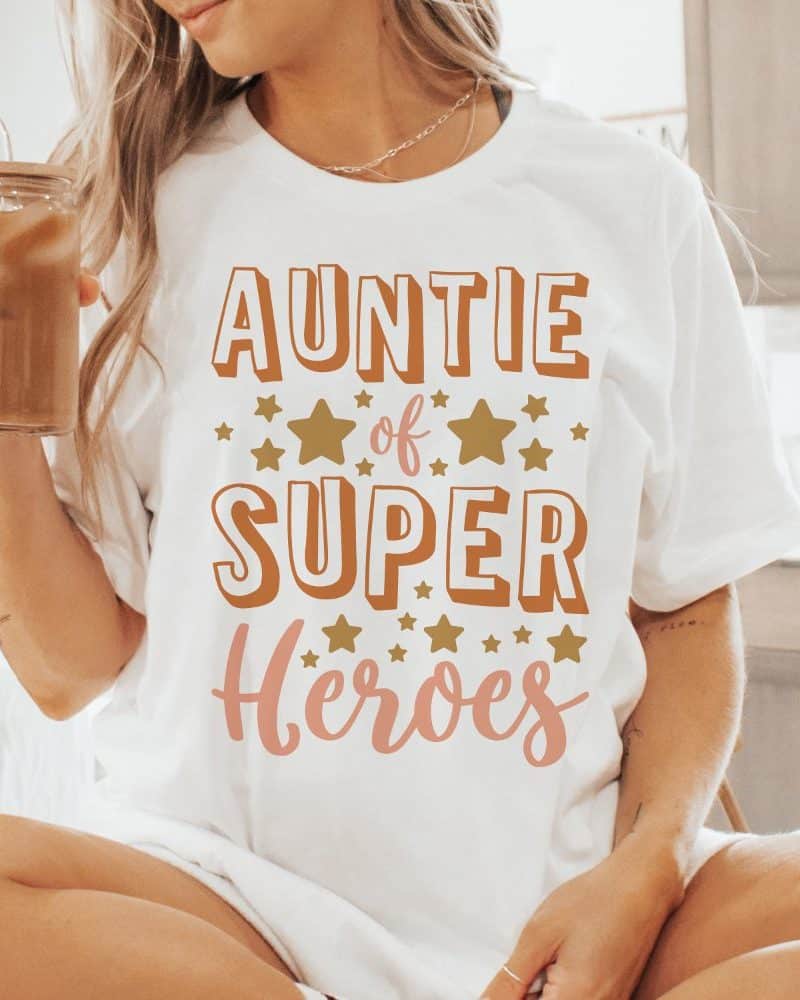 Woman wearing a white t-shirt that says Auntie of Super Heroes in orange and pink lettering with gold stars.