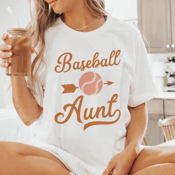 Woman Wearing A White T-Shirt That Says Baseball Aunt In Orange Script With A Baseball Graphic.