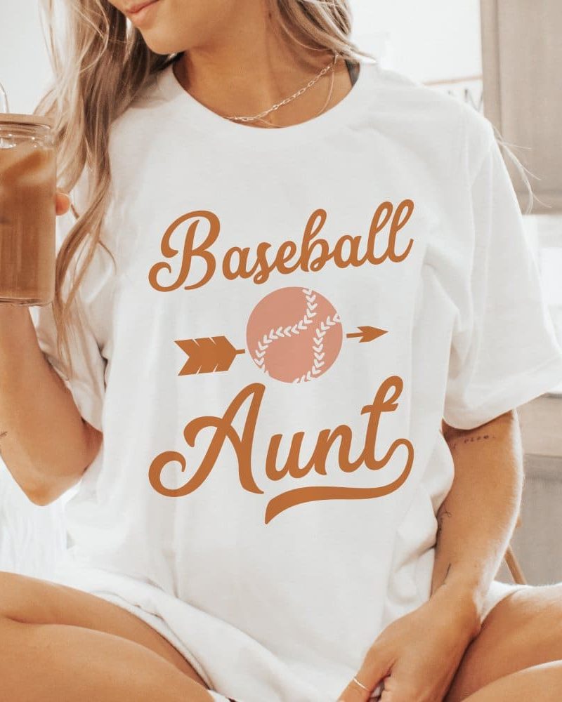 Woman wearing a white t-shirt that says Baseball Aunt in orange script with a baseball graphic.