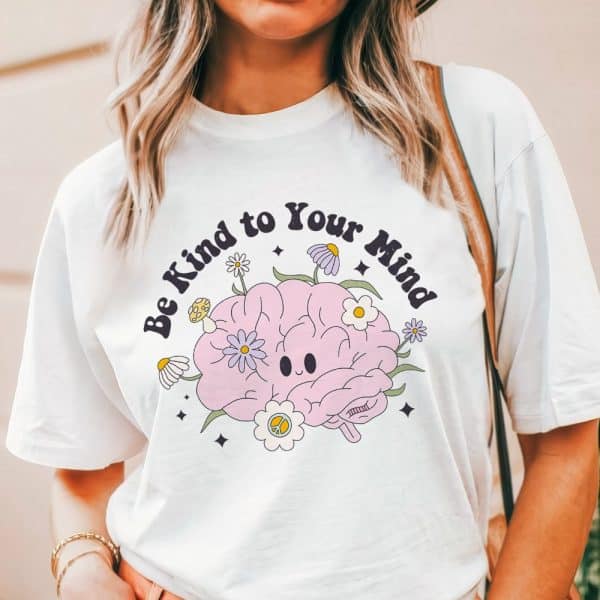 White T-Shirt With A Graphic Of A Smiling Brain Surrounded By Flowers And The Text Be Kind To Your Mind.