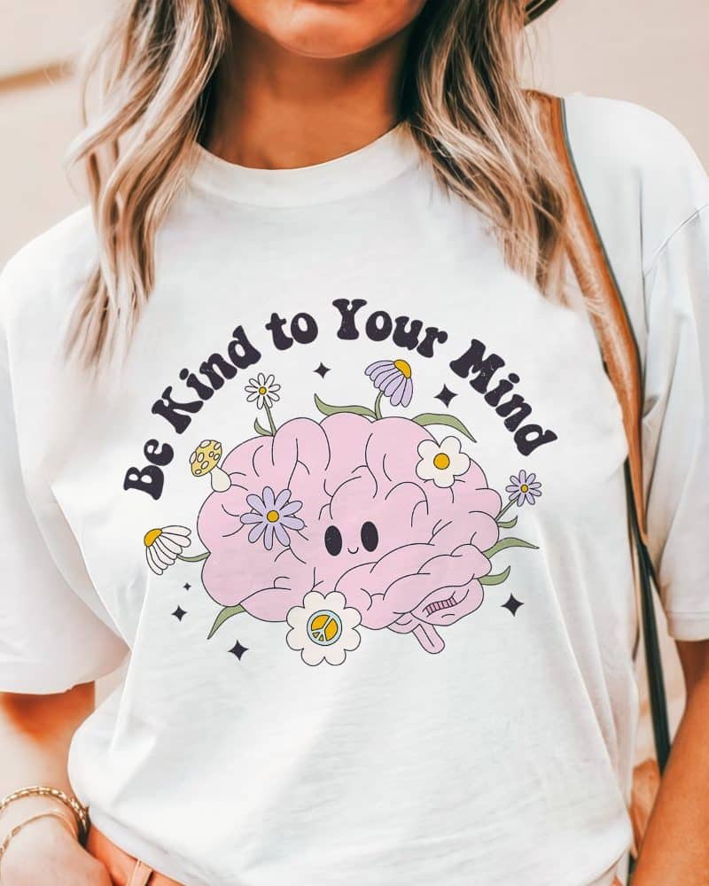 White t-shirt with a graphic of a smiling brain surrounded by flowers and the text Be Kind to Your Mind.