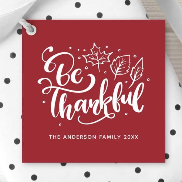 Red Gift Tag With White Lettering That Says Be Thankful, Surrounded By Small White Leaves. The Anderson Family 20Xx Is Printed At The Bottom.