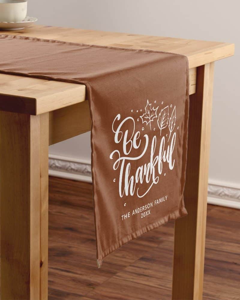 Brown table runner with white lettering that says Be Thankful, and a family name and year.
