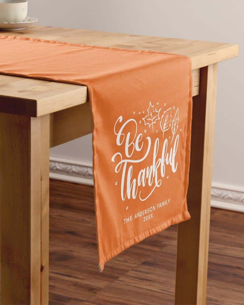 Orange table runner with the words Be Thankful in white script lettering and a small autumn leaf design.