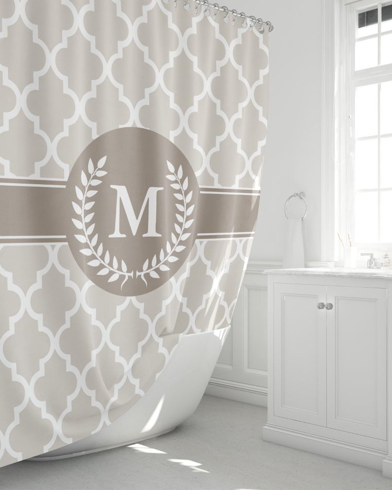 Beige shower curtain with a quatrefoil pattern and a central monogram featuring the letter M inside a laurel wreath.