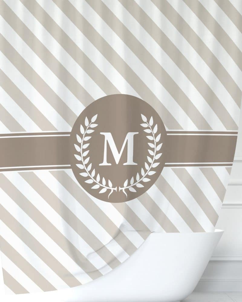 Beige and white diagonal striped shower curtain with a taupe circle and laurel wreath design centered, featuring a white capital M.