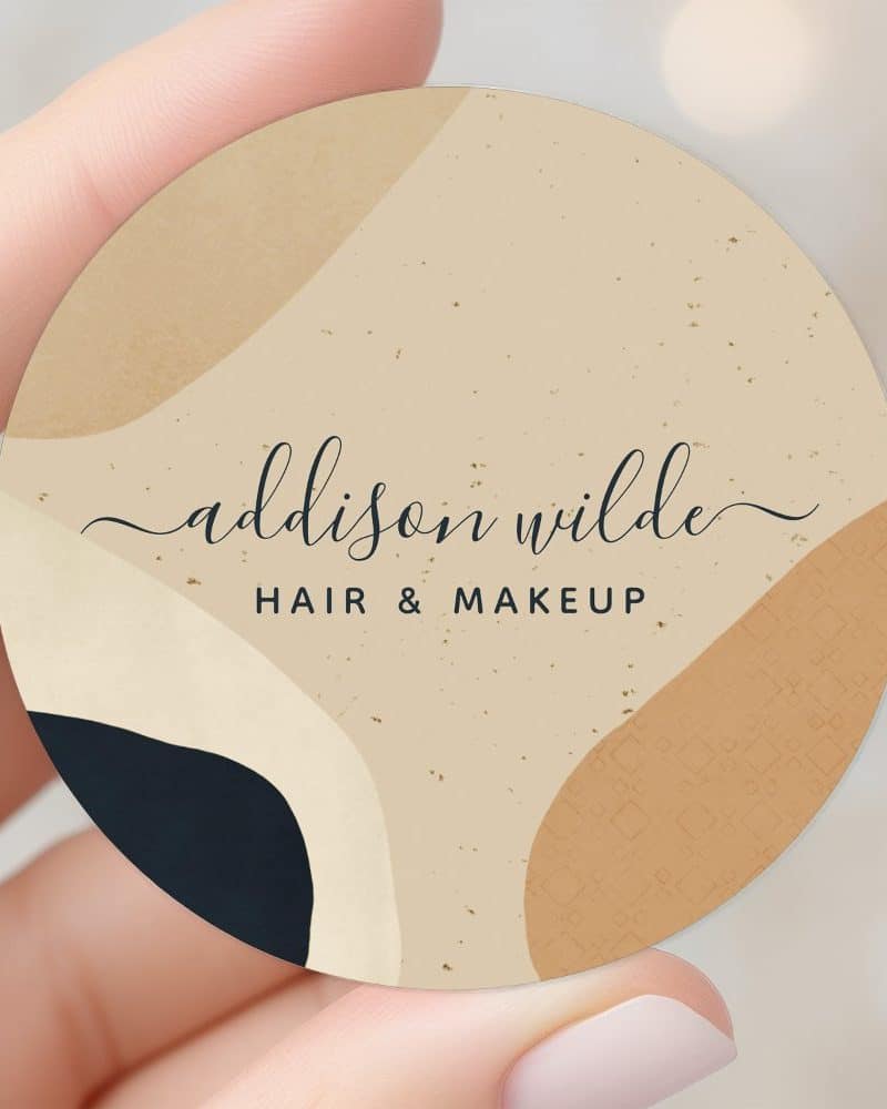 Round business card for Addison Wilde Hair & Makeup, featuring an abstract design in beige, brown, and dark blue.