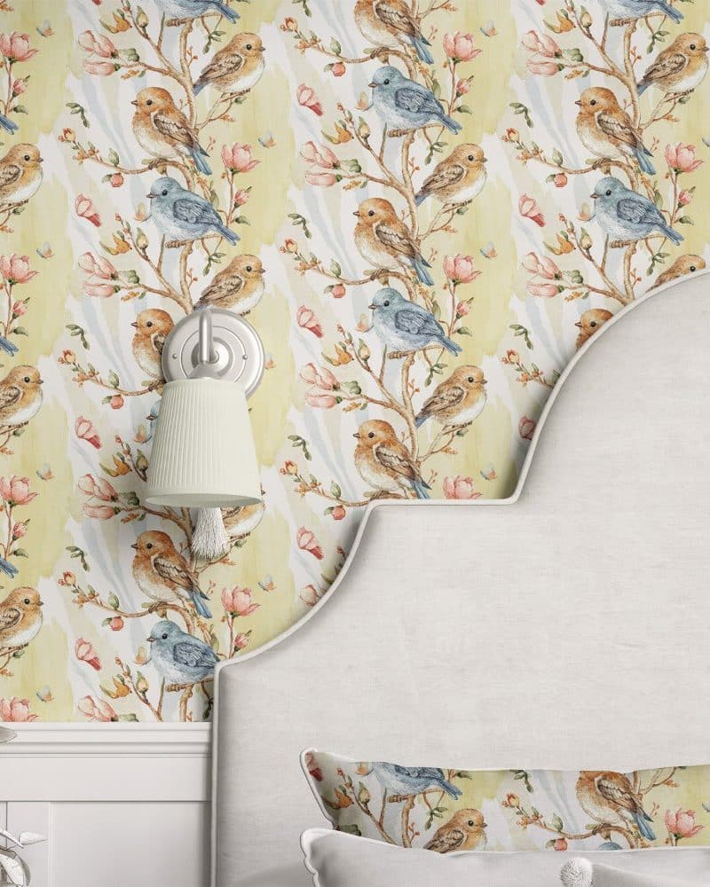 A pale yellow wallpaper featuring watercolor birds on branches with pink flowers, partially visible on a bedroom wall and headboard.