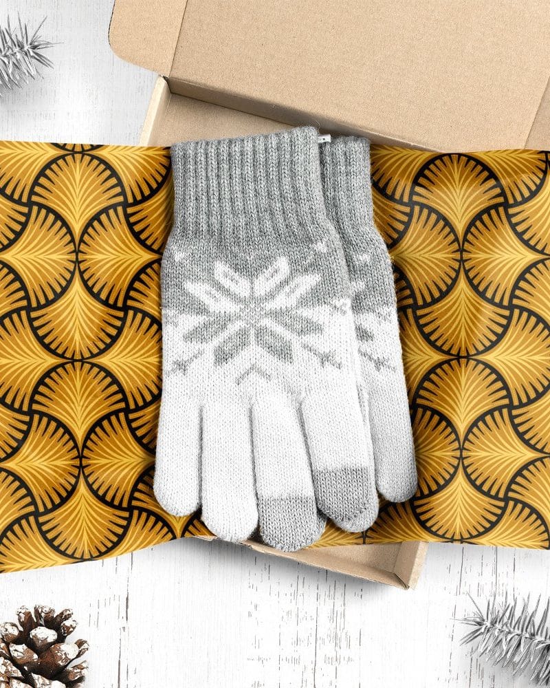 A pair of grey and white knit gloves with a snowflake pattern, inside a gift box lined with gold patterned paper.