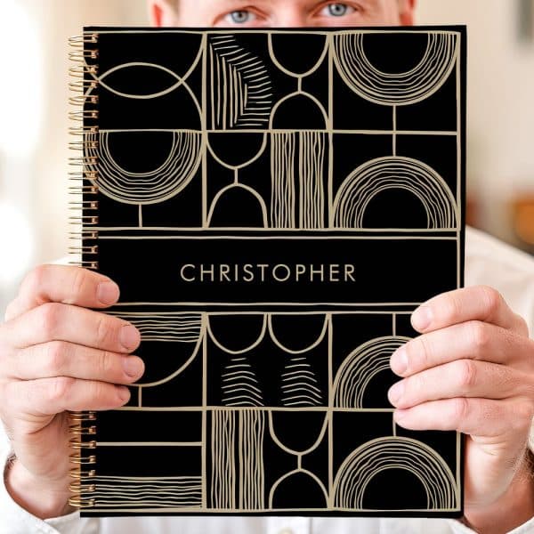 Person Holding A Black Spiral Notebook With A Gold Geometric Design And The Name Christopher On The Cover.