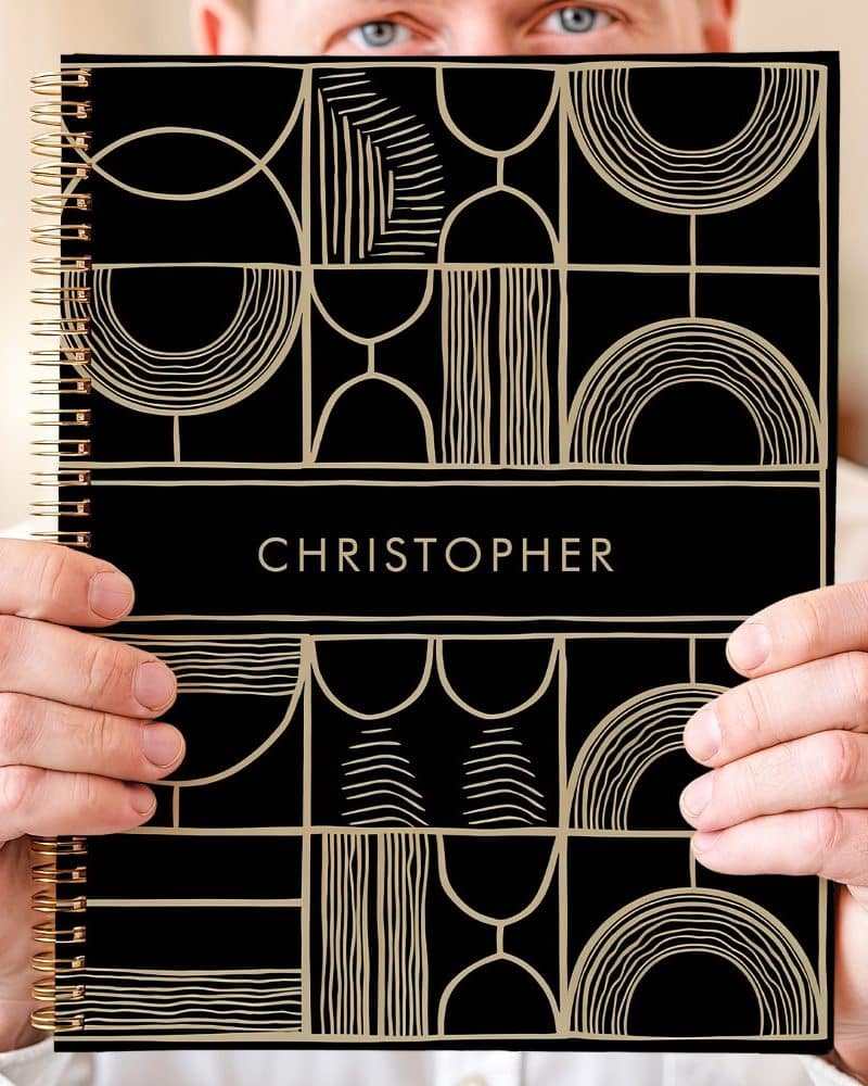 Person holding a black spiral notebook with a gold geometric design and the name Christopher on the cover.