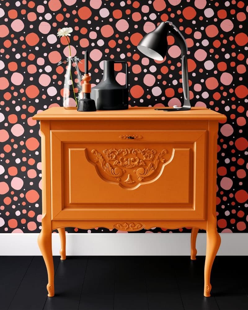 Orange painted wooden cabinet with ornate detailing, holding a lamp and decorative objects, set against a black and pink polka dot wallpaper.
