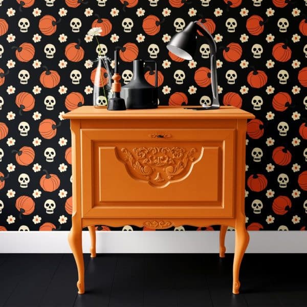 Orange Painted Wood Cabinet With Ornate Details, Decorated With A Desk Lamp And Small Vases, Set Against A Halloween Themed Wallpaper.