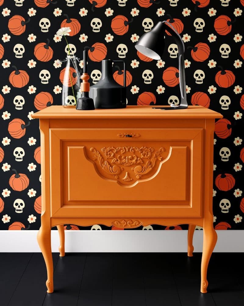 Orange painted wood cabinet with ornate details, decorated with a desk lamp and small vases, set against a Halloween themed wallpaper.