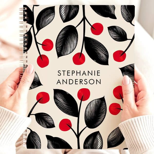 Two Hands Hold A Spiral-Bound Notebook With A Cream Background And A Repeating Pattern Of Black Leaves And Red Berries. The Name Stephanie Anderson Is Printed On The Cover.