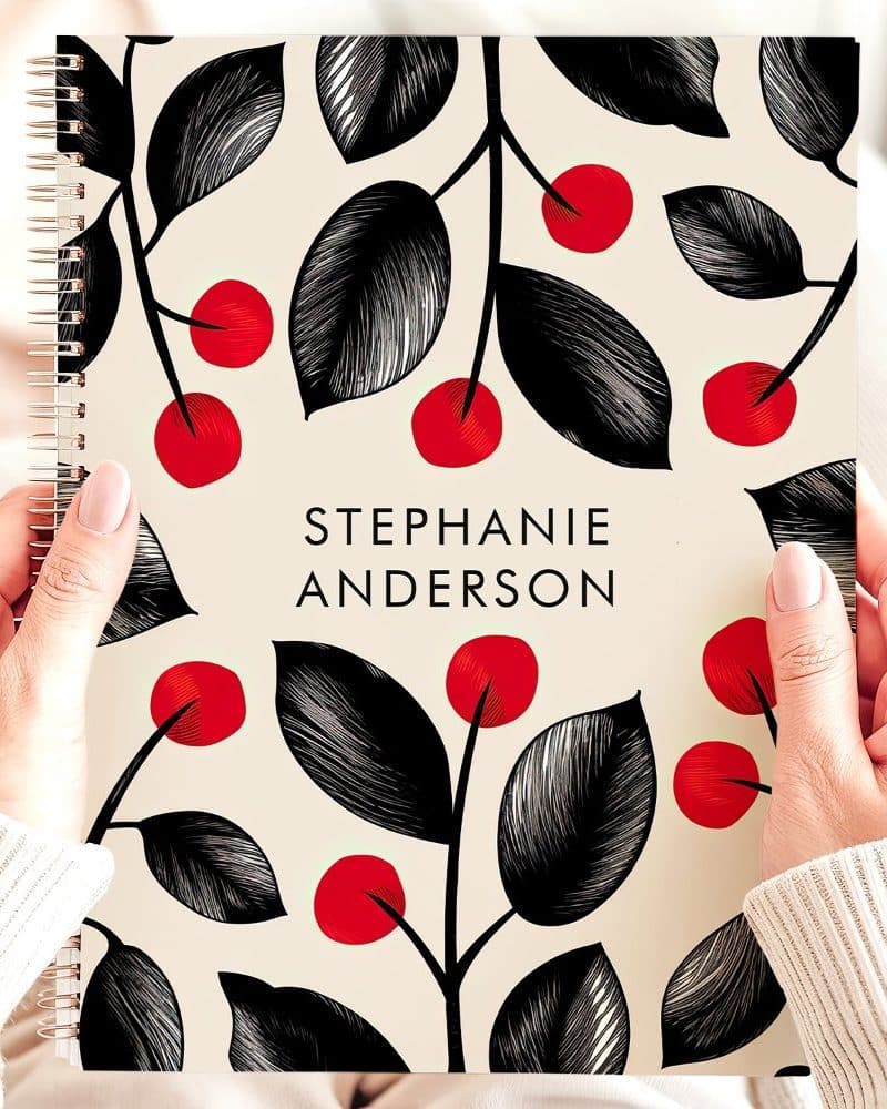 Two hands hold a spiral-bound notebook with a cream background and a repeating pattern of black leaves and red berries. The name Stephanie Anderson is printed on the cover.