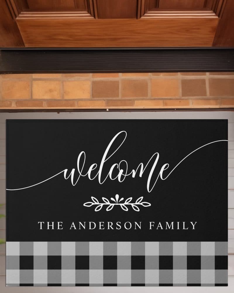Black welcome mat with white script lettering that says Welcome, a small floral design, and The Anderson Family. The bottom of the mat has a black and gray buffalo plaid design.