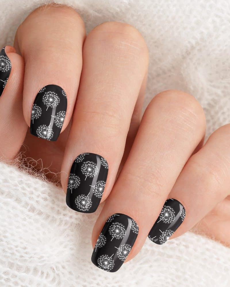 Close-up of a hand with black nails decorated with a white dandelion pattern.
