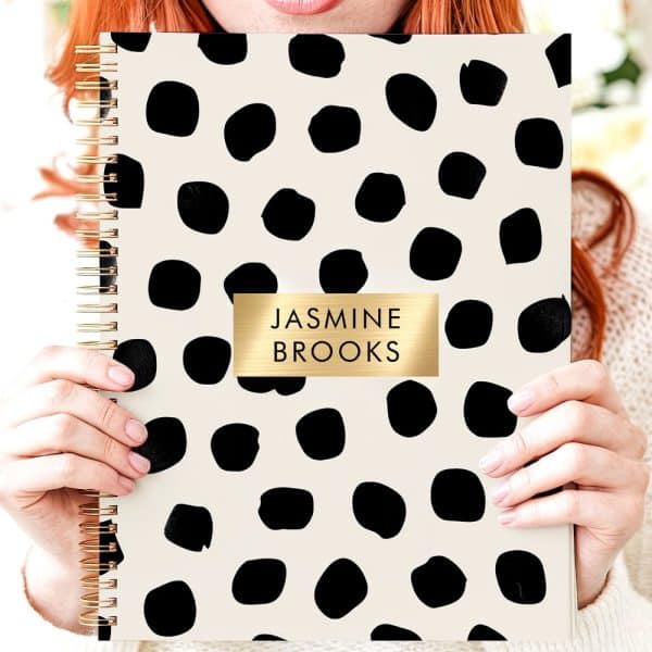 Person Holding A Cream-Colored Spiral Notebook With A Black Polka Dot Pattern And Gold Nameplate.