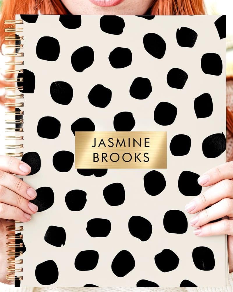 Person holding a cream-colored spiral notebook with a black polka dot pattern and gold nameplate.
