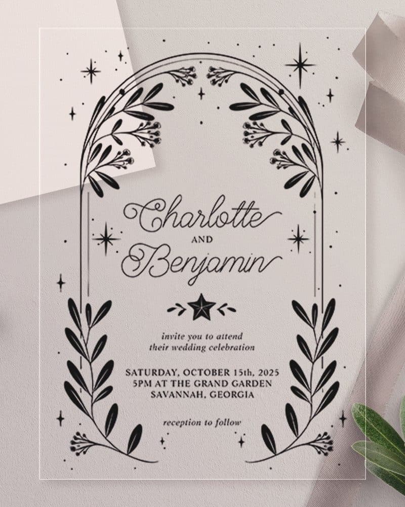 Clear wedding invitation with black floral arch and names Charlotte and Benjamin. Date and location details are included.