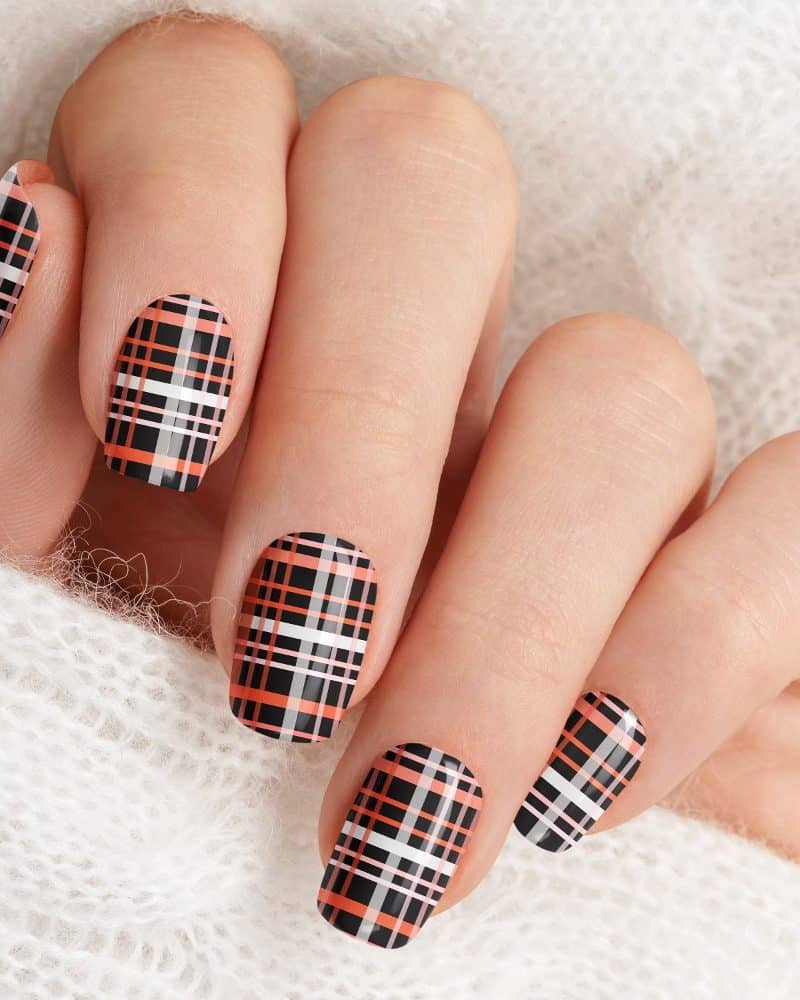 Closeup of a hand with nails painted in a black, orange, and white plaid pattern.