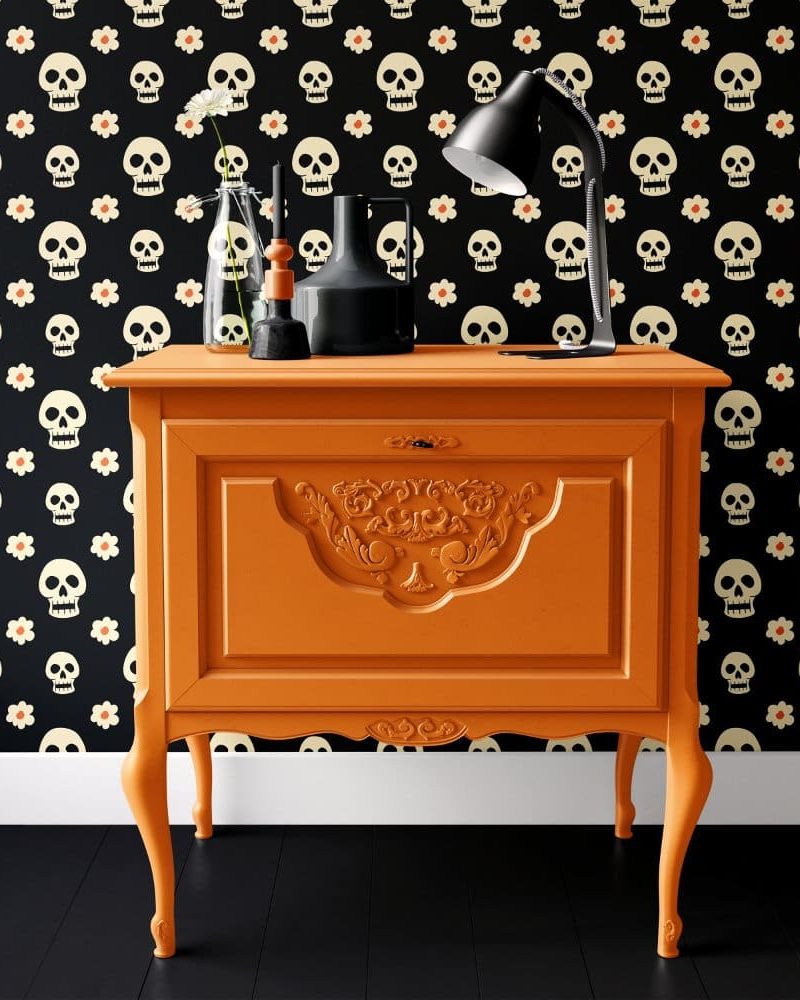 Orange wooden cabinet with ornate detailing, sitting against a wall with a skull and flower pattern wallpaper. A desk lamp and decorative objects are on top.