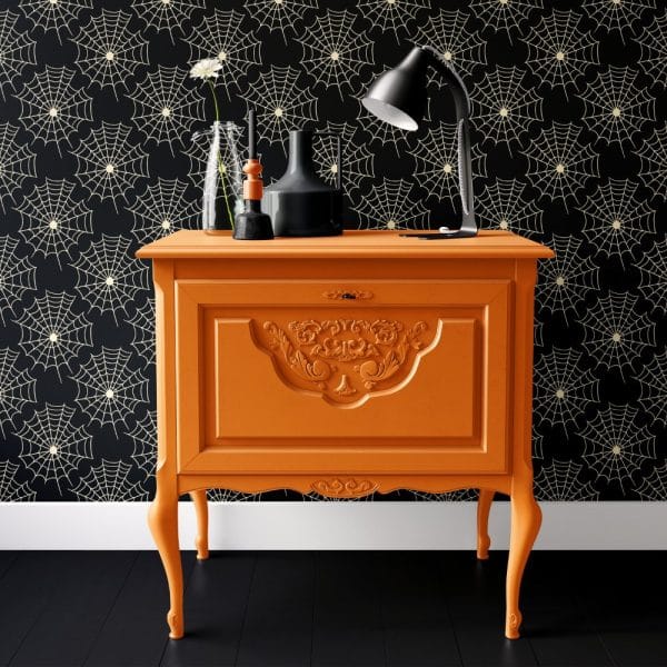 An Orange Painted Antique-Style Cabinet Sits Against A Black Wall With A Cream-Colored Spiderweb Pattern.