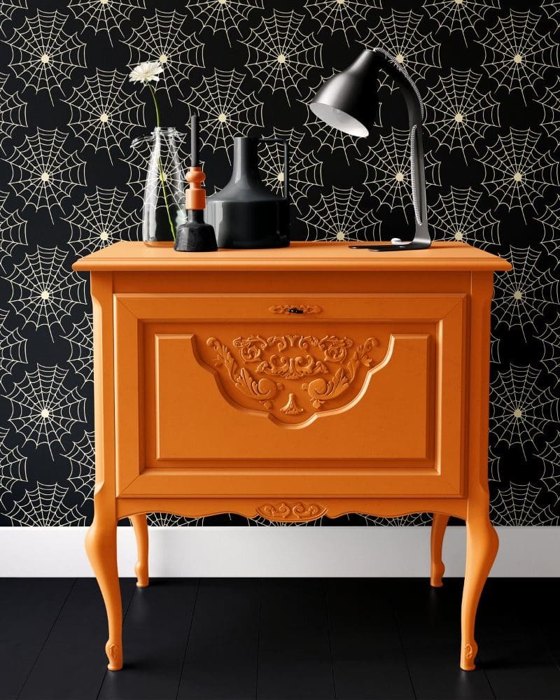 An orange painted antique-style cabinet sits against a black wall with a cream-colored spiderweb pattern.
