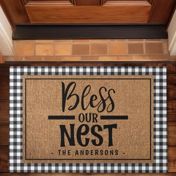 A Tan Doormat With Black Lettering That Says Bless Our Nest, And The Andersons Below. The Doormat Has A Black And White Checkered Border.