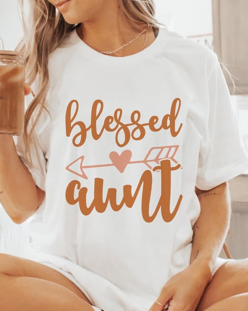 Woman wearing a white t-shirt that says Blessed Aunt in brown lettering with an arrow and heart graphic.