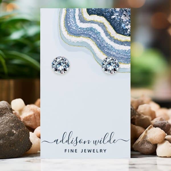 Pair Of Round, Clear Gemstone Stud Earrings Displayed On A Light Blue Jewelry Card.