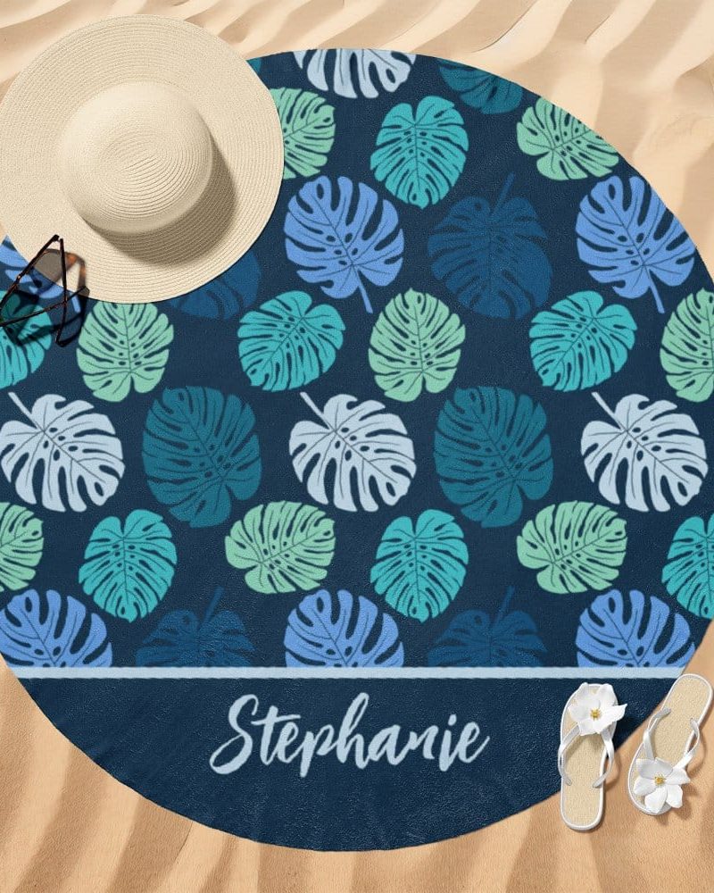 Round beach towel on sand with a dark blue background, teal and light blue monstera leaf pattern, and the name Stephanie at the bottom.
