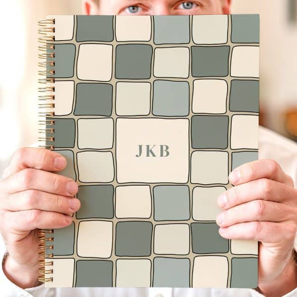 A Person Holds Up A Spiral-Bound Notebook With A Beige And Green Tile Pattern And The Initials Jkb In The Center.