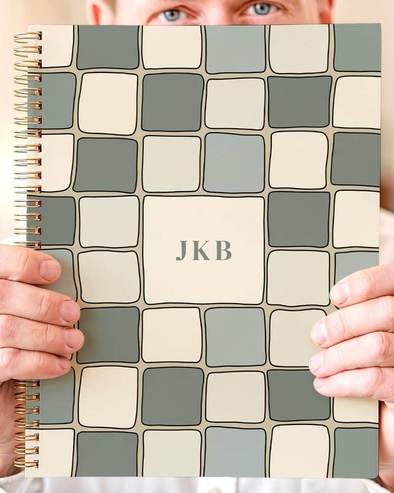 A person holds up a spiral-bound notebook with a beige and green tile pattern and the initials JKB in the center.