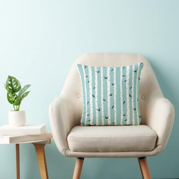 A Light Beige Armchair With A Teal And White Patterned Throw Pillow Featuring Small Birds And Tree Trunks.