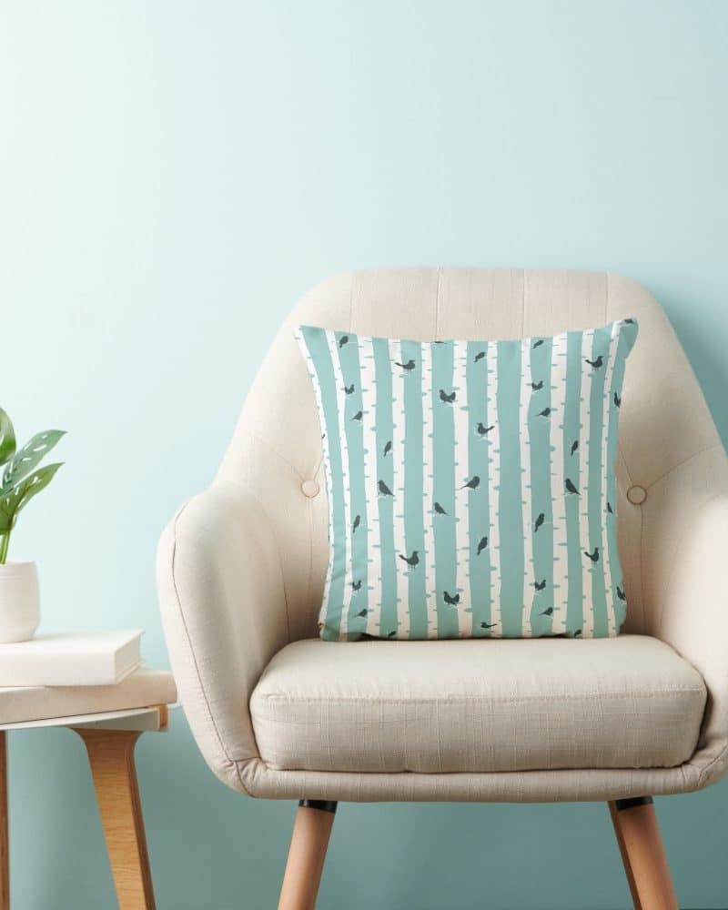 A light beige armchair with a teal and white patterned throw pillow featuring small birds and tree trunks.
