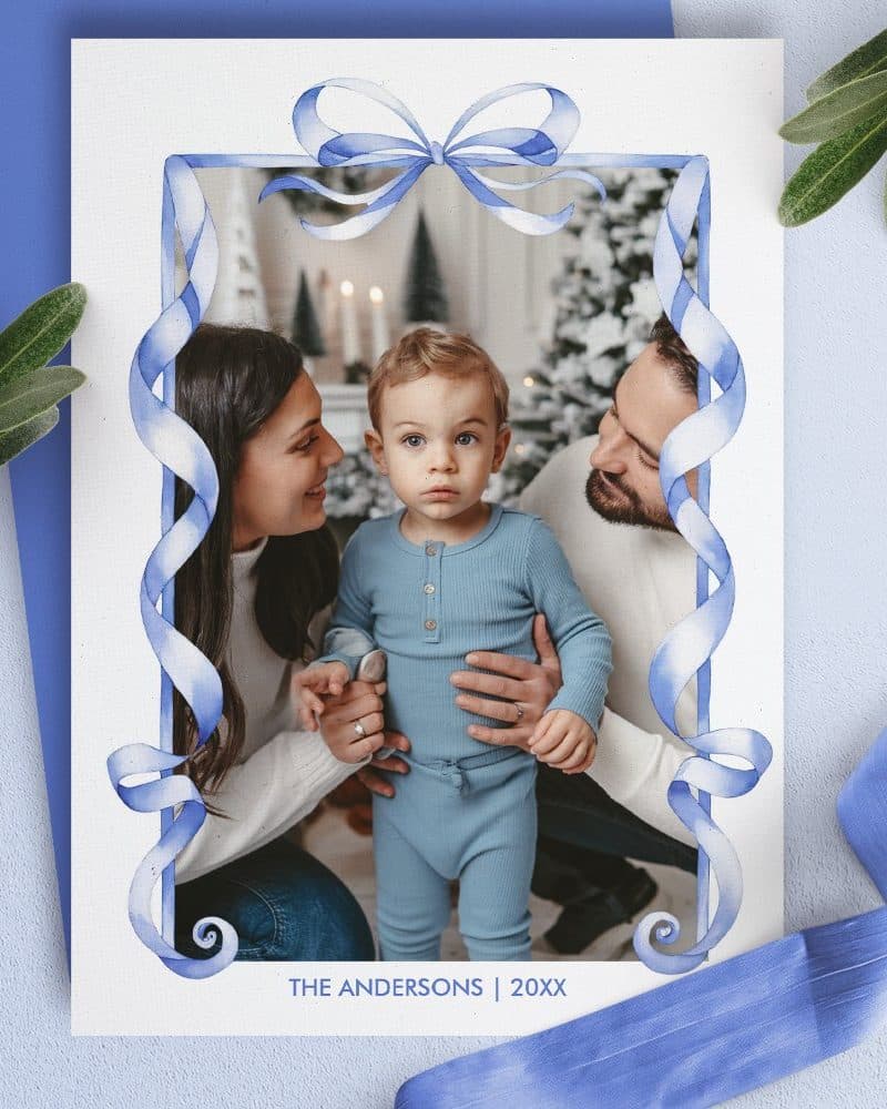 Christmas card featuring a family photo of parents and their toddler child, framed by a watercolor blue ribbon border.
