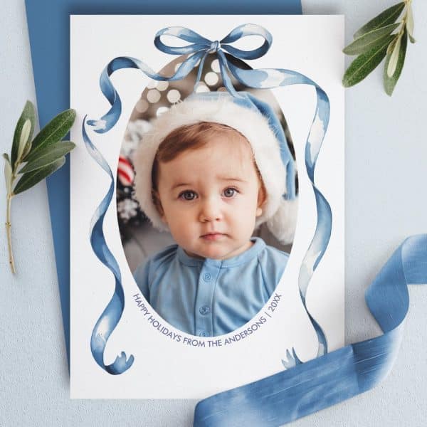 A Holiday Card Featuring A Baby Wearing A Santa Hat In An Oval Frame Decorated With Blue Watercolor Ribbons And A Bow. The Andersons' Holiday Greeting Is At The Bottom.