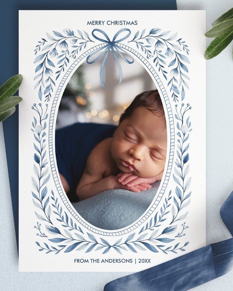 Christmas card featuring a sleeping baby in an oval frame decorated with blue watercolor foliage.